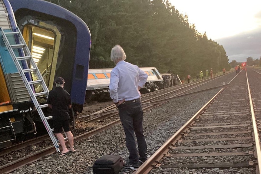 A train lies on its side, a man with his hands on his hips looks at the carriage on its side.