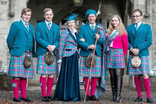 A family stands wearing tartan kilts and green jackets outside an old building.
