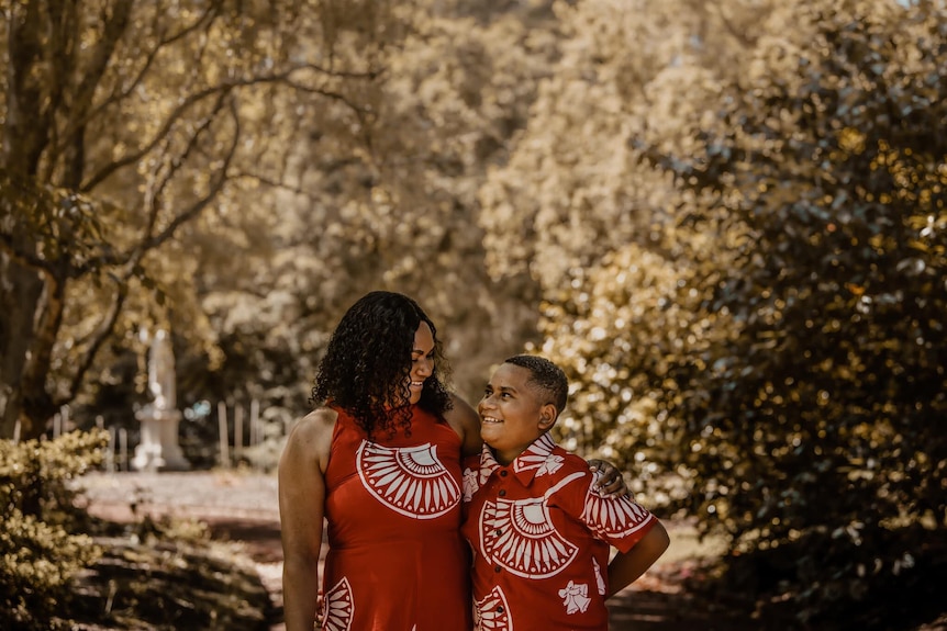 Woman and son wear red and white matching outfits. Smiling at each-other, surrounded by parkland. 