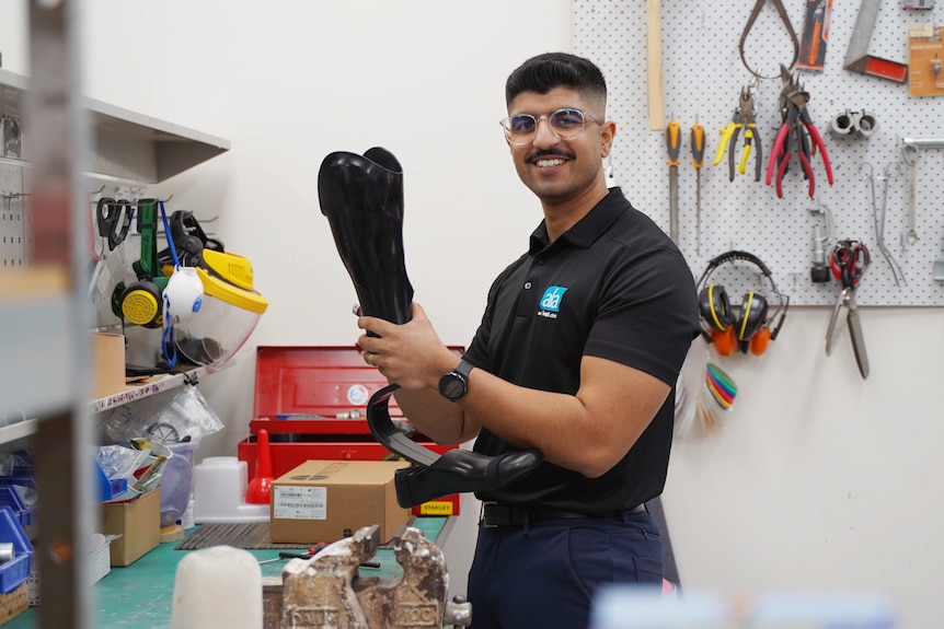 Man hold up prosthetic limb whilst smiling at the camera, he stands at a workstation surrounded by tools.