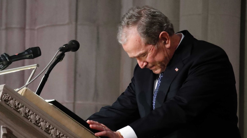 Former US President George W Bush bows his head while standing at a lectern.