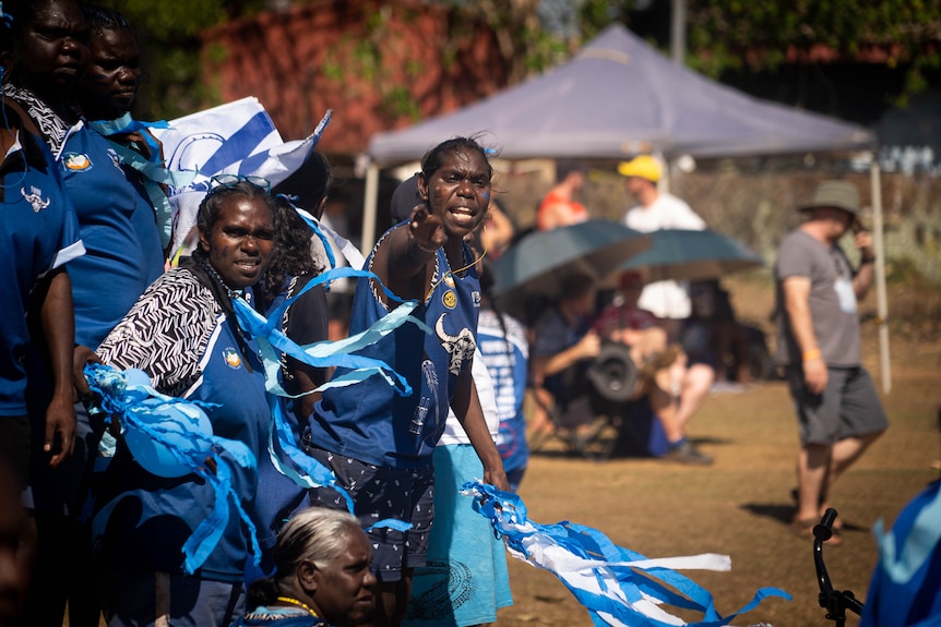 Young women in blue jersies hold blue streamers and watch a football game which is out of shot