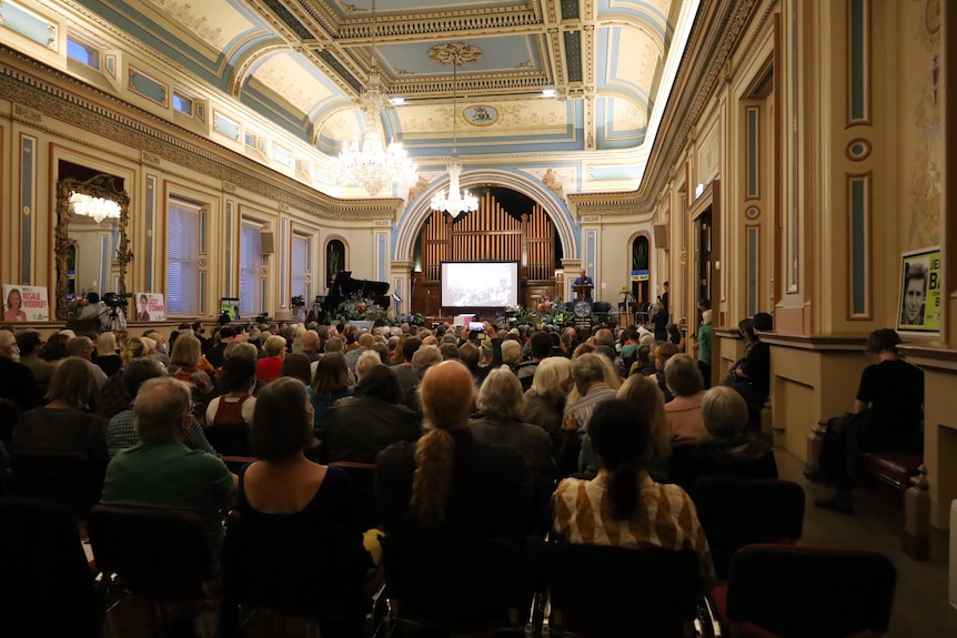 A hall full of people taken from the back, looking towards the stage where a man is speaking