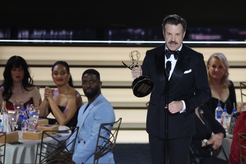 Jason Sudeikis sosteniendo un Emmy y vistiendo un traje en el escenario