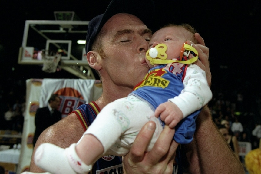 A basketball player kissing a baby sucking on a dummy and wearing an Adelaide 36ers jersey