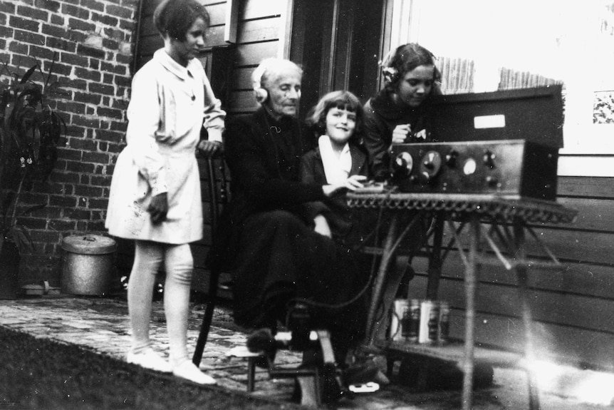 A black and white photo of a woman using Alfred Traeger's pedal radio, with three children watching on.