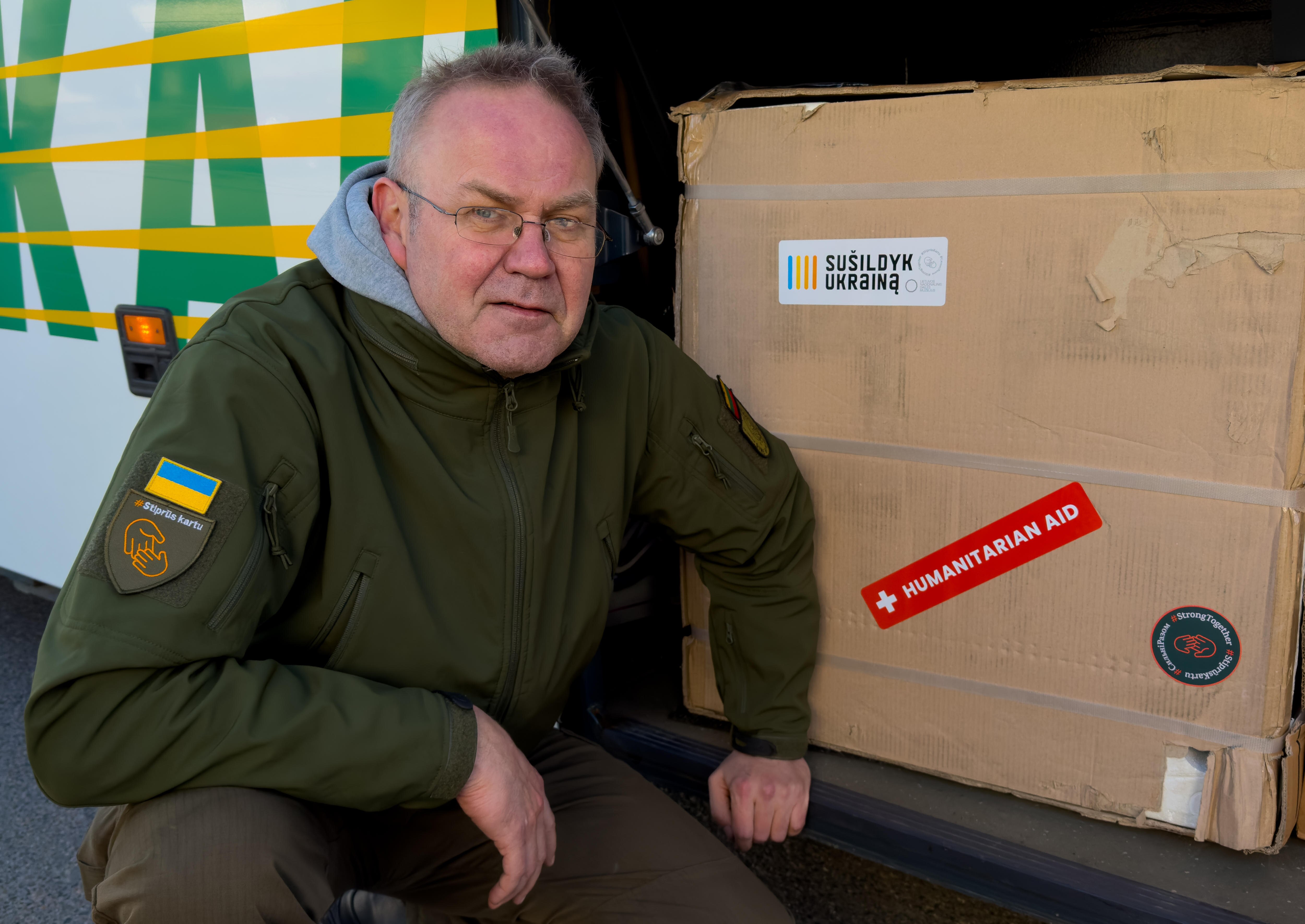 A man in glasses wearing a khaki jacket crouches by a box labelled 'humanitarian aid'