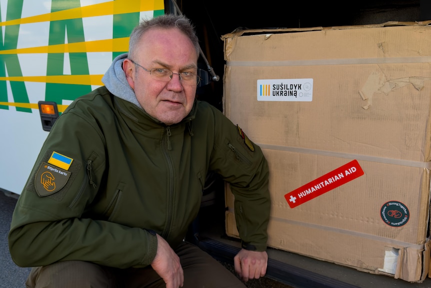 A man in glasses wearing a khaki jacket crouches by a box labelled 'humanitarian aid'