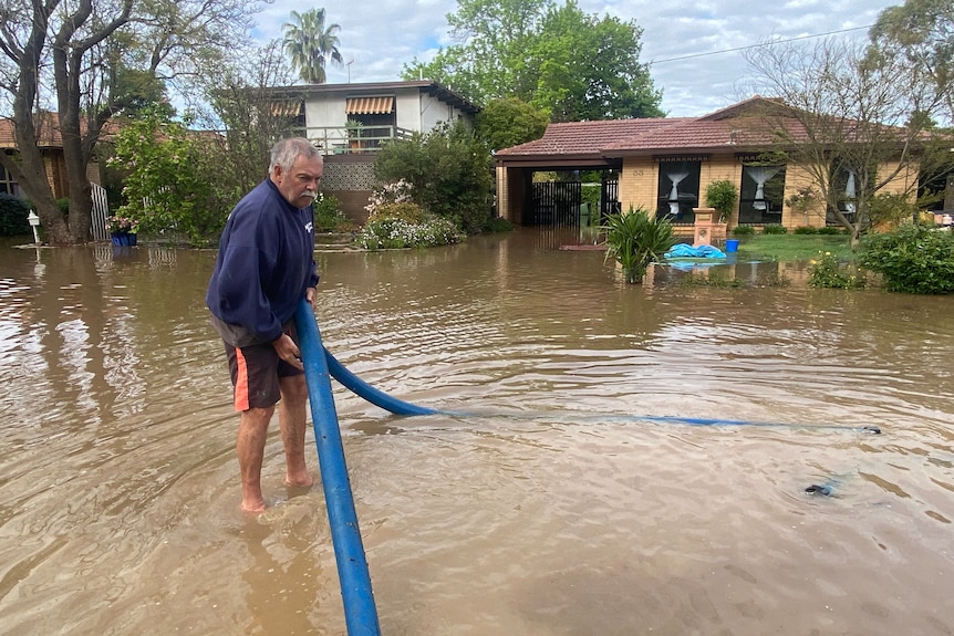 A man stands in ankle-deep flood water as he holds up a large blue pipe that is submerged underground. 