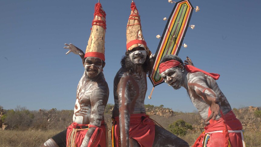 Three Aboriginal dancers stand posed on a beach