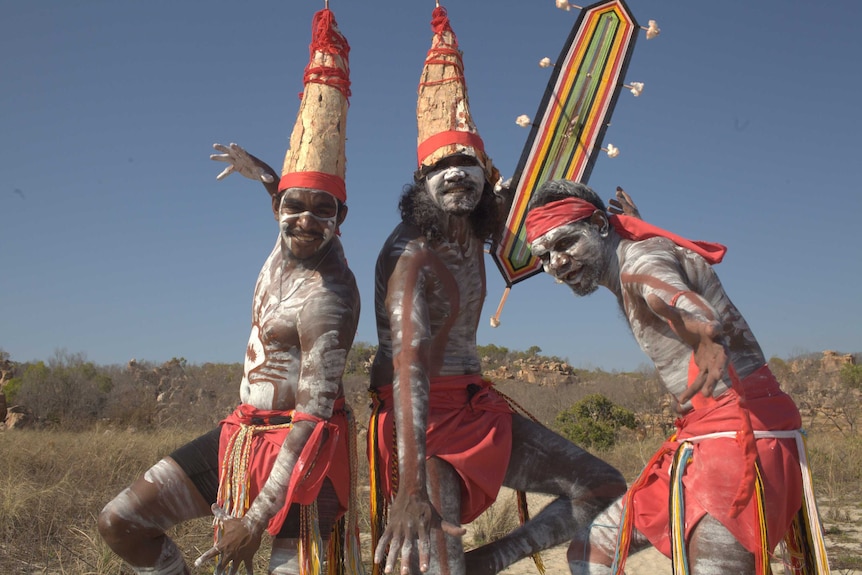 Three Aboriginal dancers stand posed on a beach