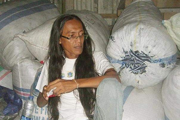 A man with long hair sits against bags of recycling. 