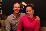 A man and woman in their thirties sit side-by-side at a table. She has a red dress on and he has a button up shirt.