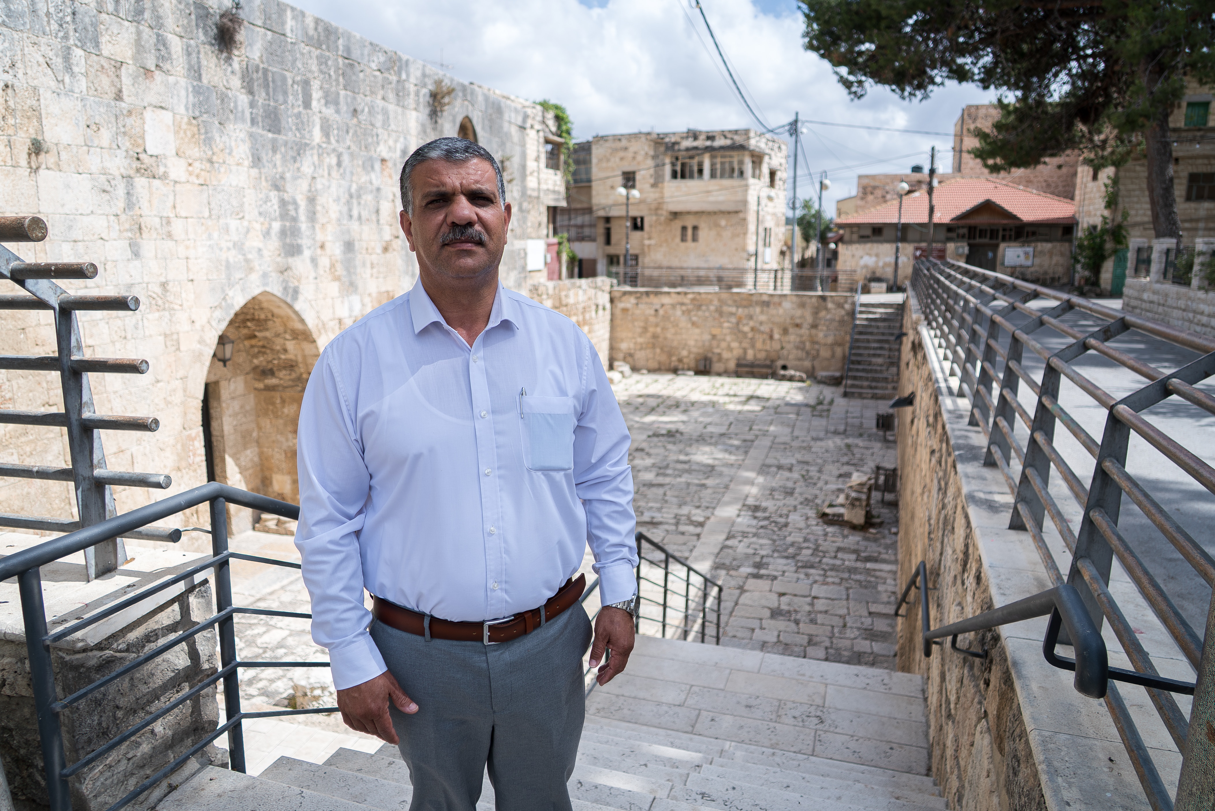 A Palestinian man wearing a shirt and pants stands with his arms by his sides.