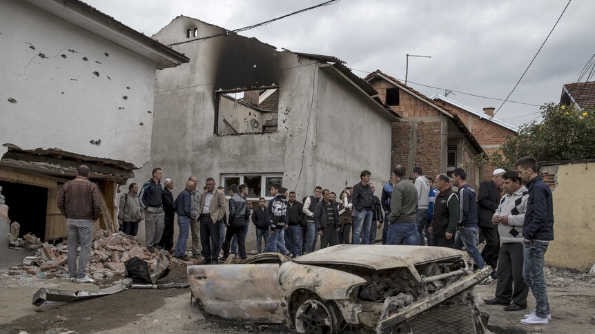 Residents stand beside a burnt out vehicle in Kumanovo