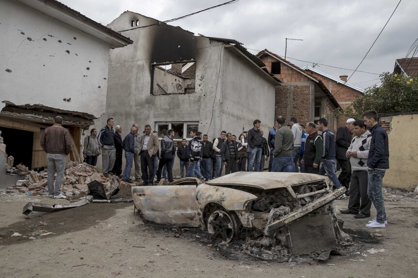 Residents stand beside a burnt out vehicle in Kumanovo
