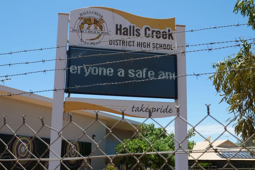 The sign for Halls Creek District High School behind a barbed wire fence.