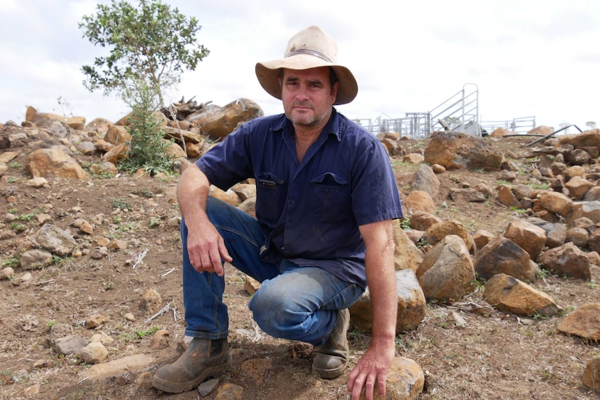 A middle aged man kneels in a rocky dirt field. He's wearing a beaten Akubra, work shirt and jeans