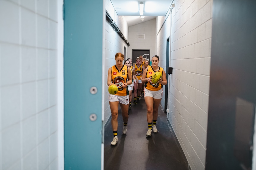 A women's Aussie rules team walks through a corridor before a game, with two players holding footballs.