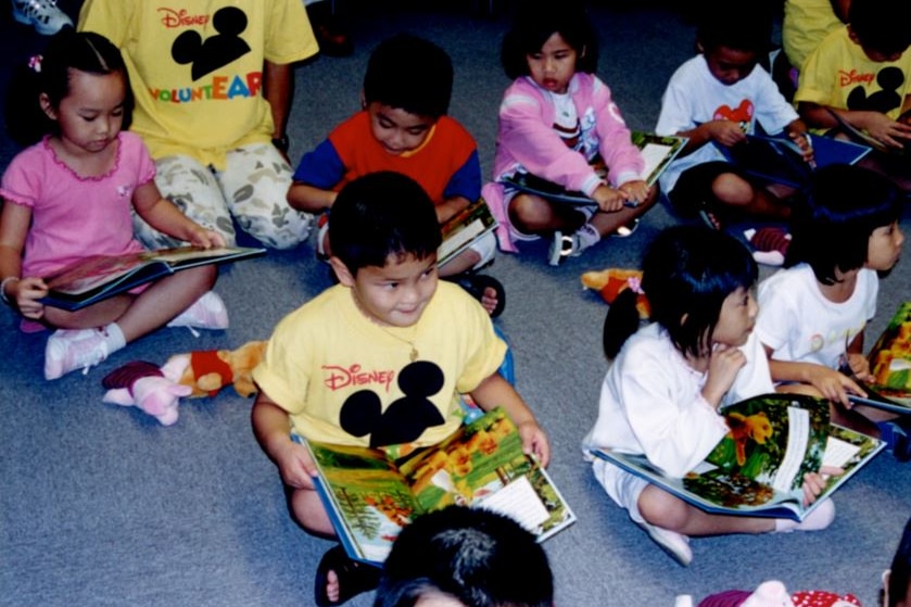 An Asian child in a yellow t-shirt sits at the front of two rows of children with a book open on his lap. He looks to the front.