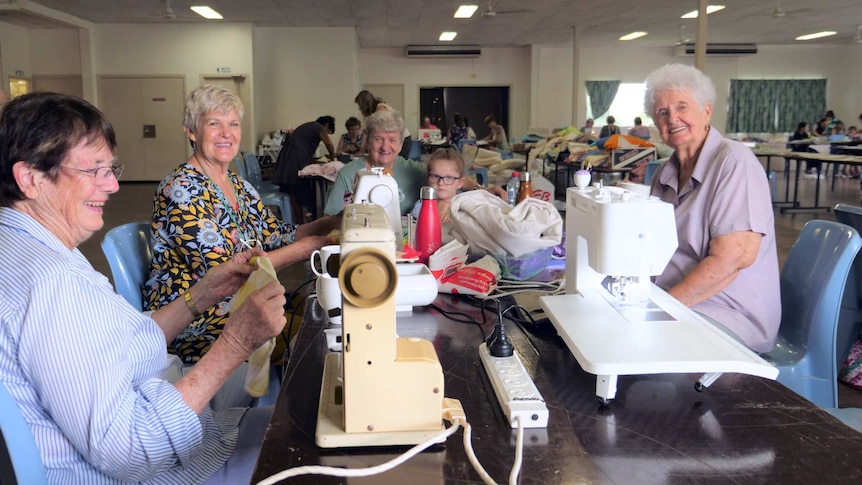 Four older women and a girl sit around sewing machines in a hall.