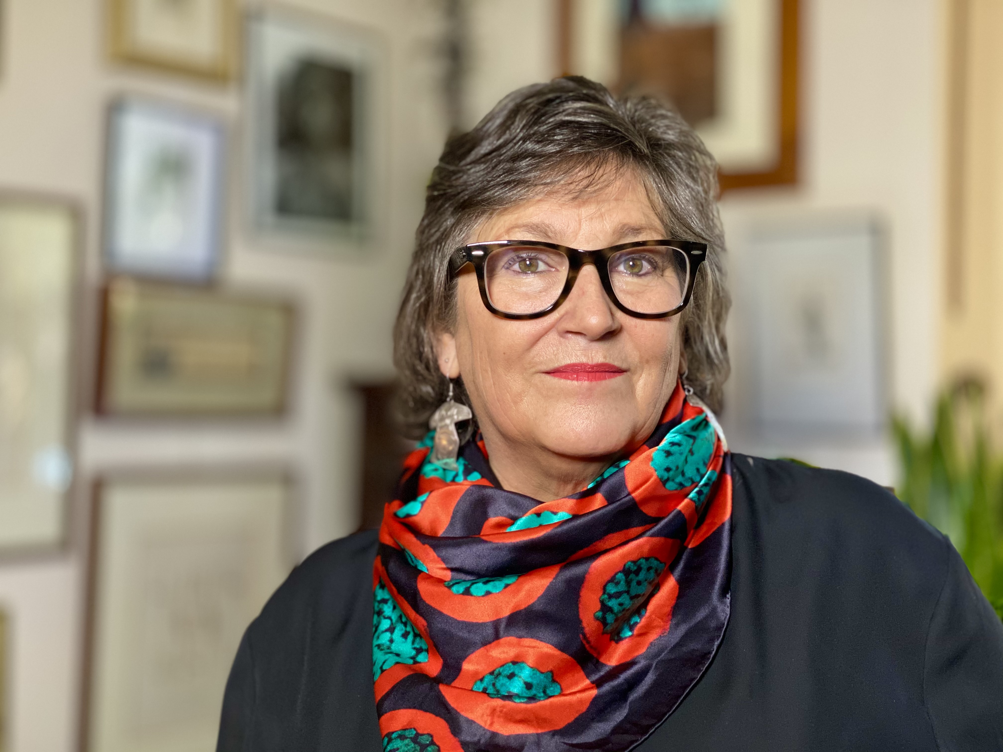 A woman wearing a black shirt and brightly coloured scarf and glasses looks at the camera