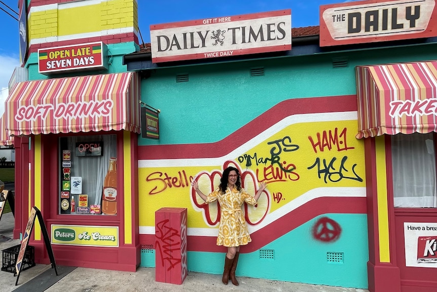 A woman stands outside a brightly coloured corner store. 