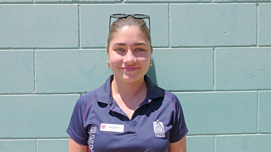 A young girl wearing a navy polo shirt and sunnies on her head stands infront of a pale blue wall smiling at camera