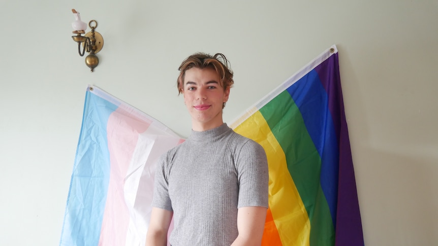Transgender woman standing in front of queer and transgender flags in grey t-shirt smiling