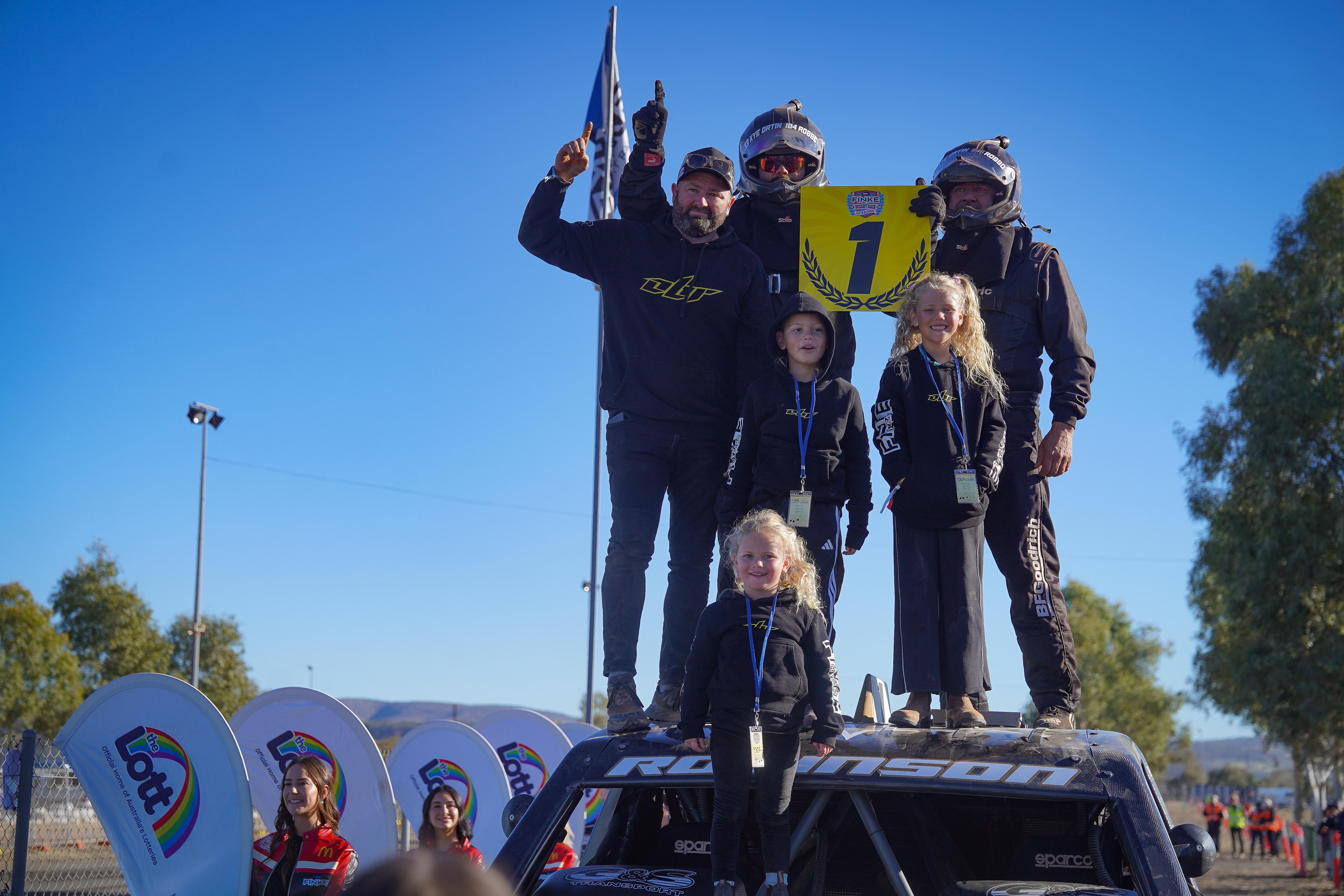 A small group of racing car drivers and their young family members standing on top of a car and holding a sign saying "1".