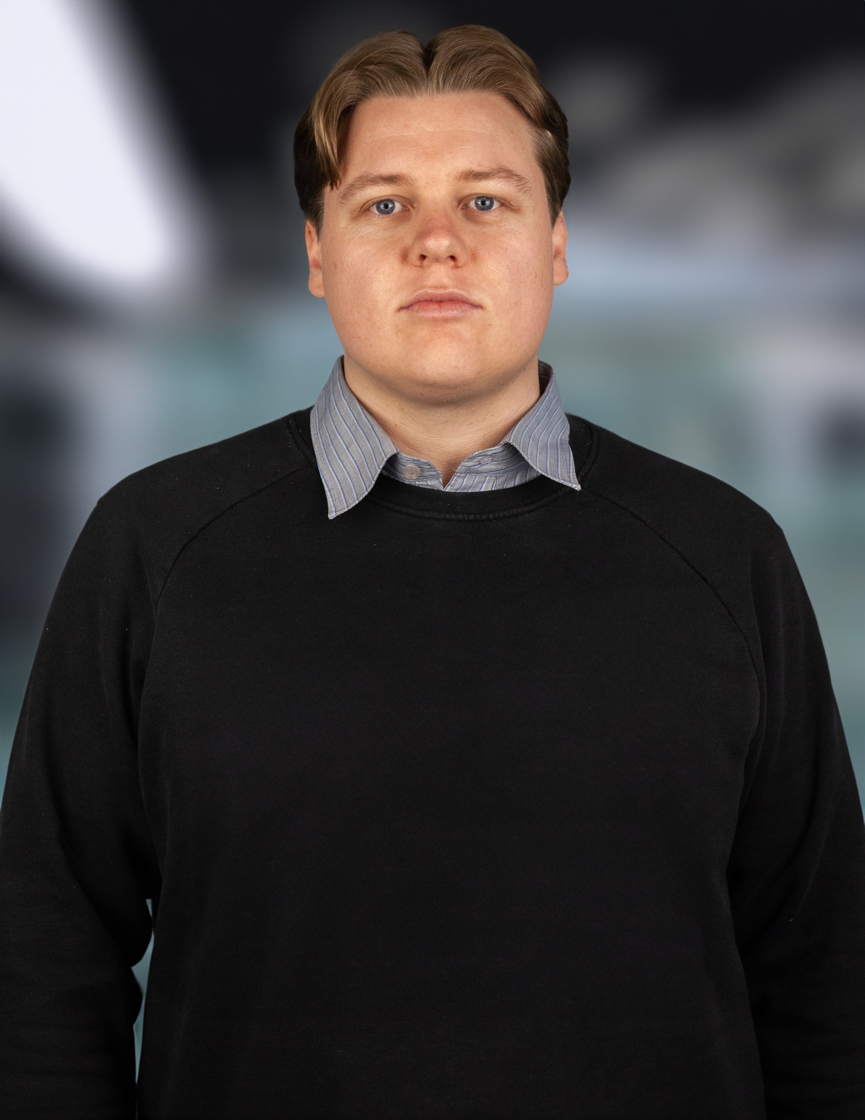 A man stands looking at the camera wearing a dark sweater and collared shirt. The background is out of focus.
