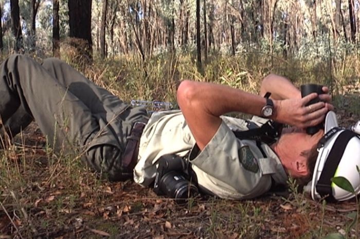 Man in green uniforms lies on grass, face up, looking upwards using binoculars.