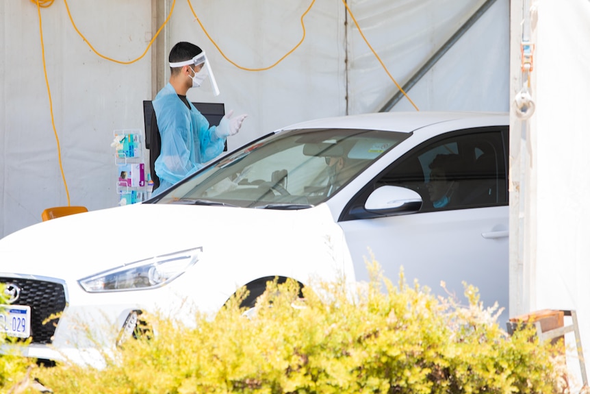 A man in blue PPE standing near a white car under a tent.