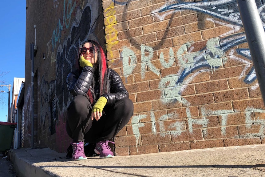 A woman with dyed pink haid crouches in front of graffiti