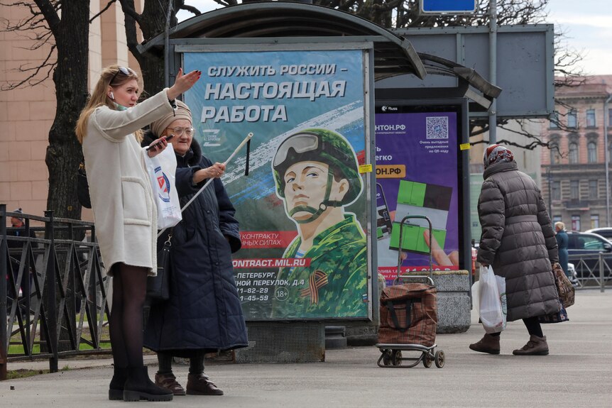 A young woman and an elderly lady stand in front of a bus stop the features a big poster in Russian with a soldier illustrated.