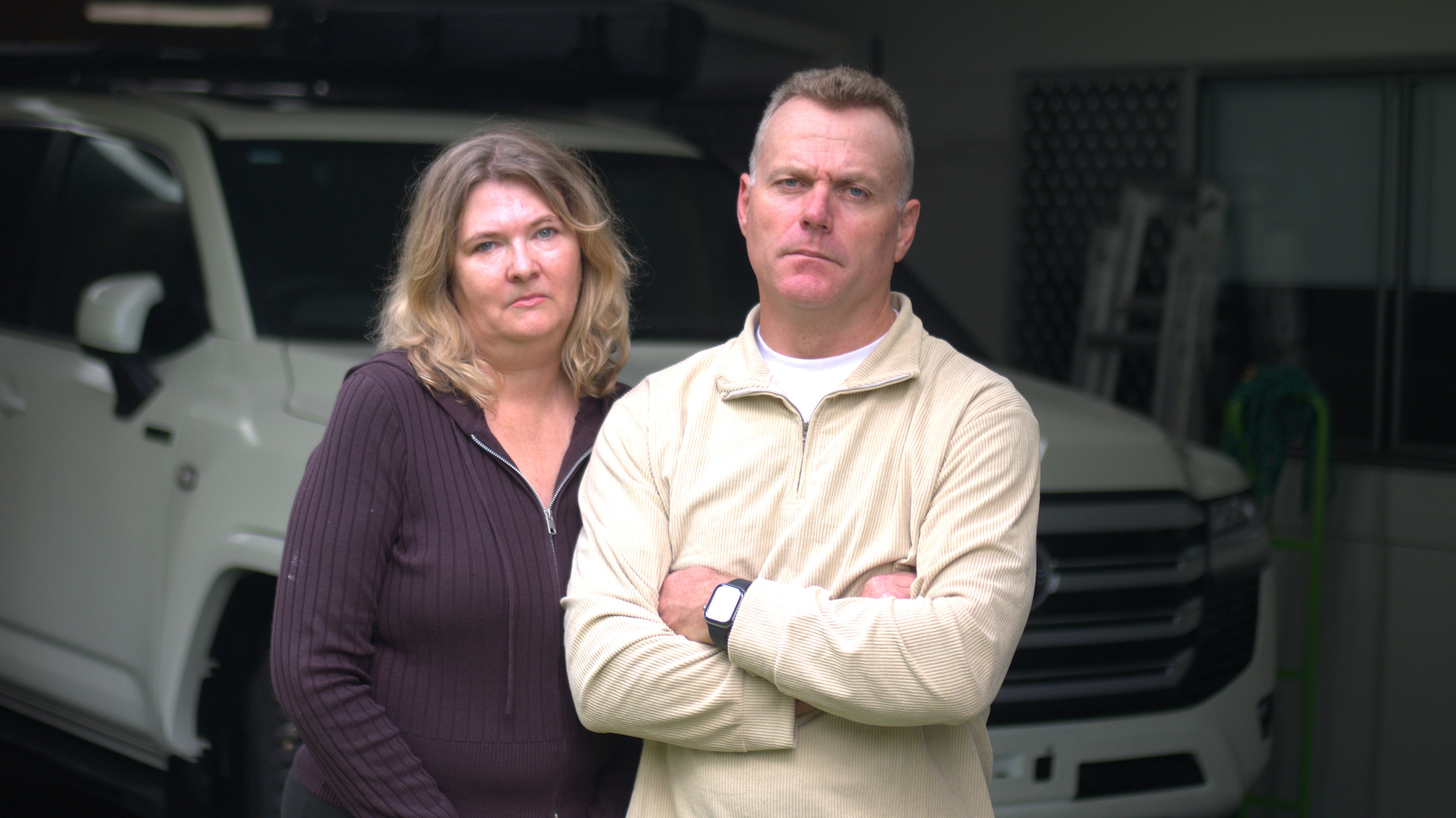 woman and man standing cross-armed and frowning in front of car