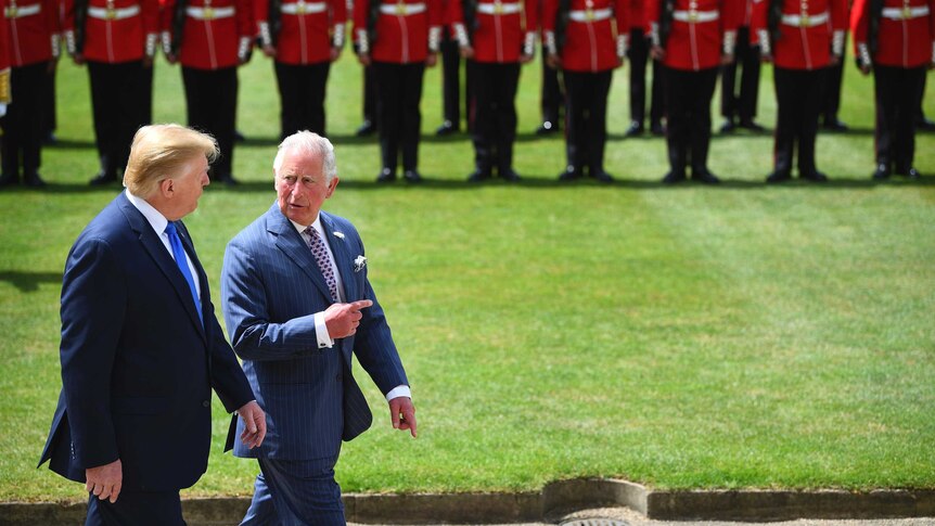Charles points ahead as he walks with Trump in front of a line of British royal guards in red with black hats.