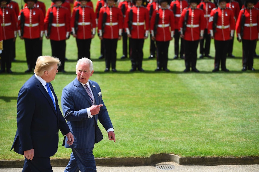 Charles points ahead as he walks with Trump in front of a line of British royal guards in red with black hats.