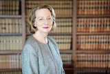 Portrait of a woman in her late sixties who is in front of a book case. She has short hair and light blue eyes.