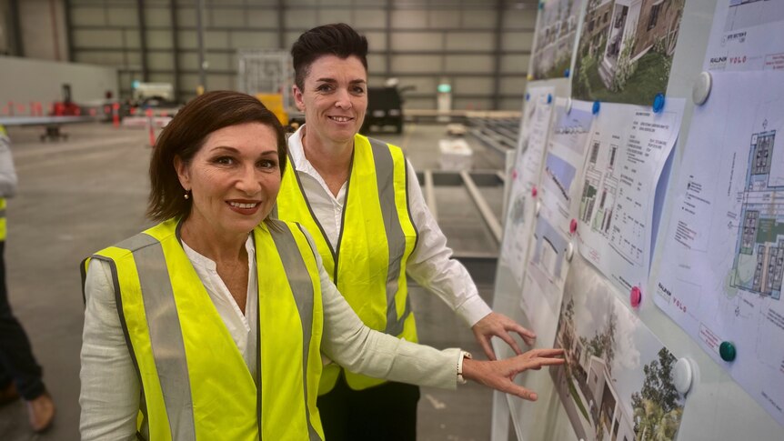 two politicians wearing high-vis vests over their clothes point to a wall with various building plans on it, smiling