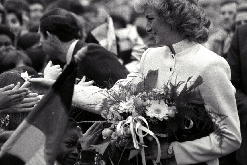 A black and white close up of Princess Diana meeting wellwishers while holding flowers with Prince Charles standing beside her.