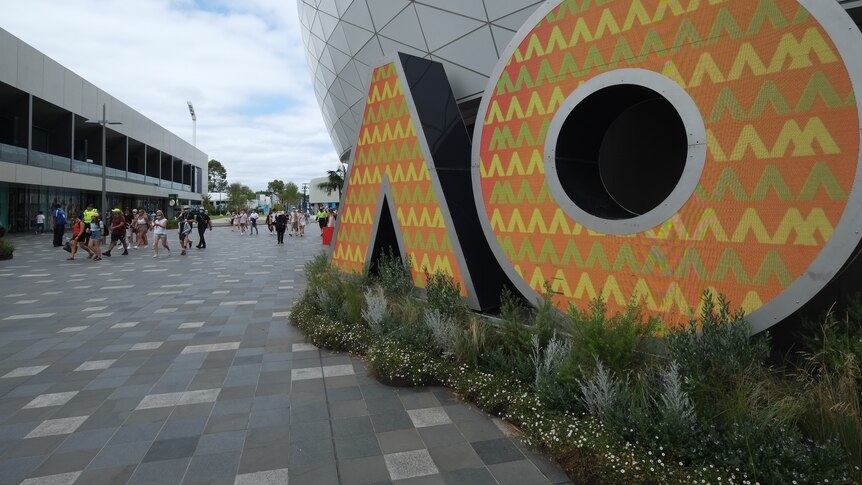 Giant yellow AO sign in front of white tessellated building, with crowds in the distance to left of frame.