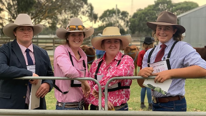 Four young agriculture students dressed in hats and brightly coloured shirts lean on a farm fence. 