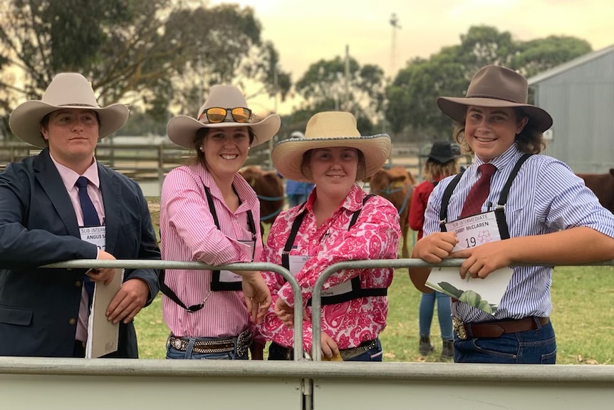 Four young agriculture students dressed in hats and brightly coloured shirts lean on a farm fence.