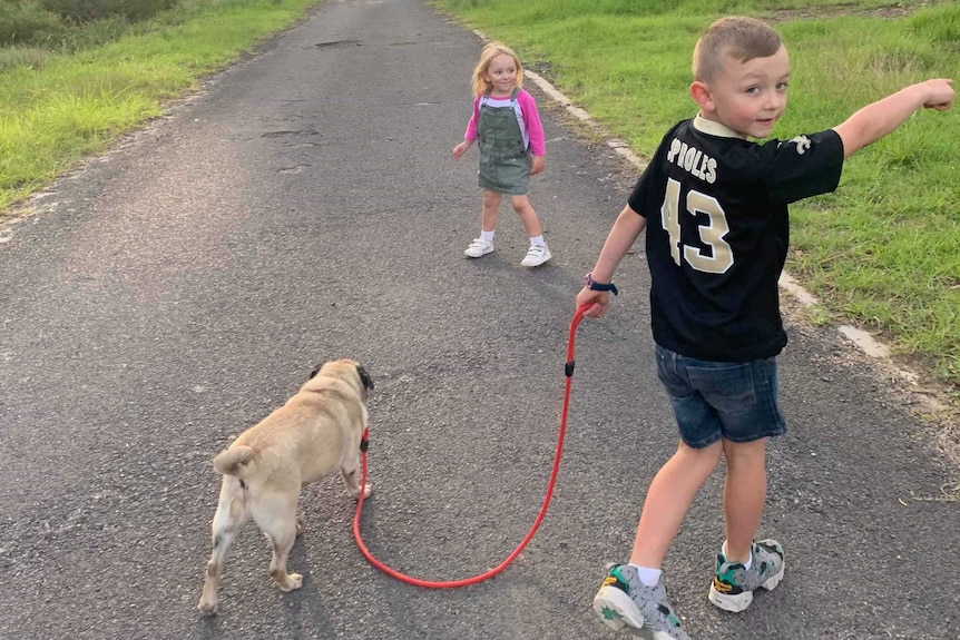 Two children walk down a country road with a small dog on a lead.