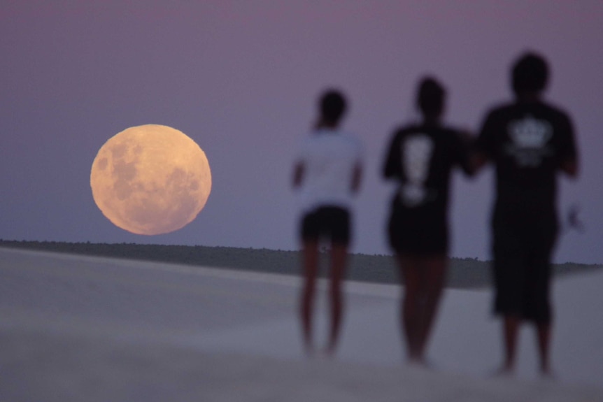 A giant moon on the horizon, with three people silhouetted next to it.