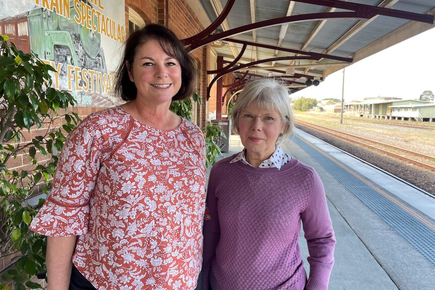 Two women stand smiling on a country railway station.