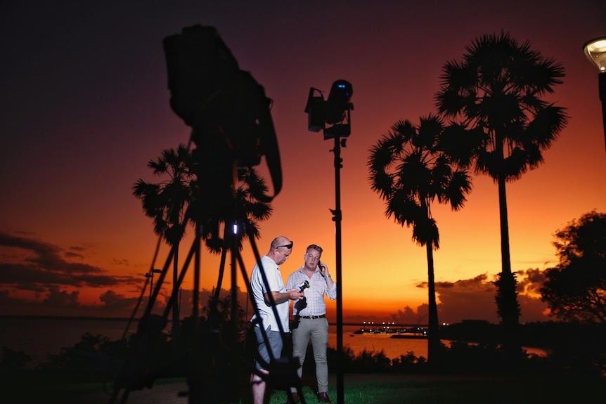 Journalist and cameraman standing under palm tree, light stand and in front of camera, silouhetted against red sunrise sky. 