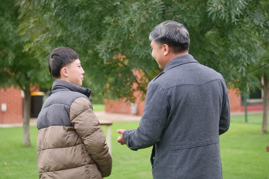 A teenage boy wearing a puffer jacket speaks with a man. Both have their backs to the camera.