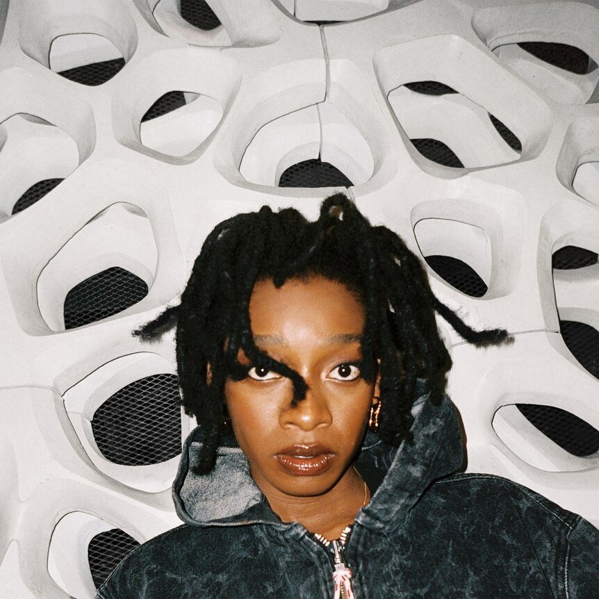 a woman with dreadlocks in a blue tracksuit top seated in front of a wall of speakers.